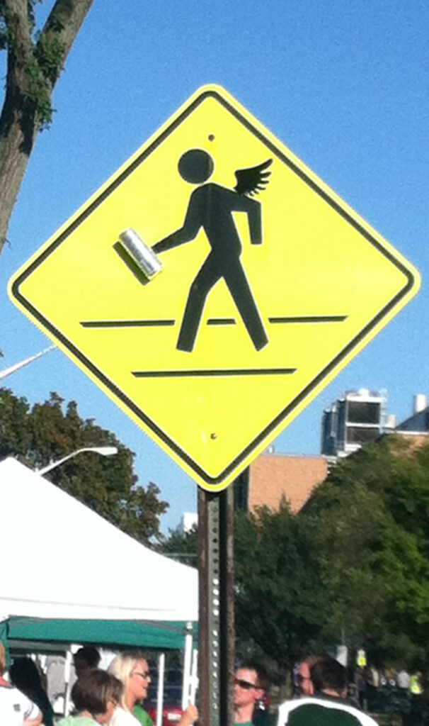 A pedestrian crossing sign with a pair of wings and a Red Bull can added on.