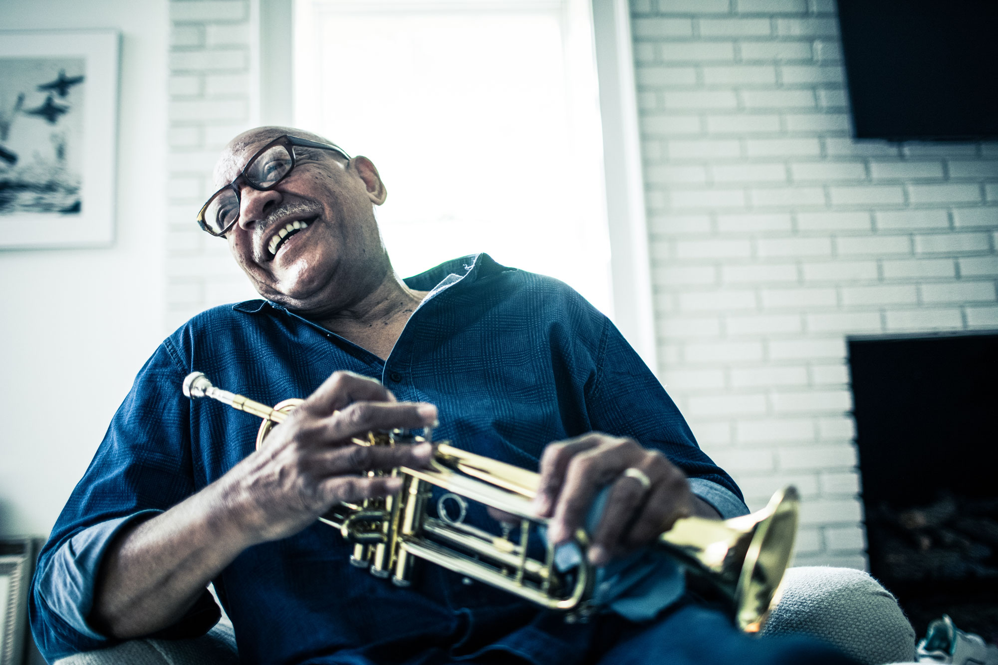 An older man sits on a chair, his head tilted back in laughter, a trumpet in his hands