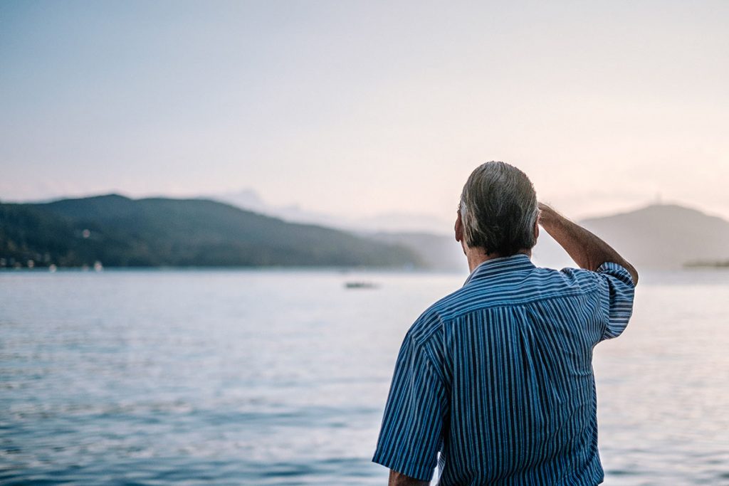 A gray-haired man looks out over a lake, hills beyond, his right hand shading his brow