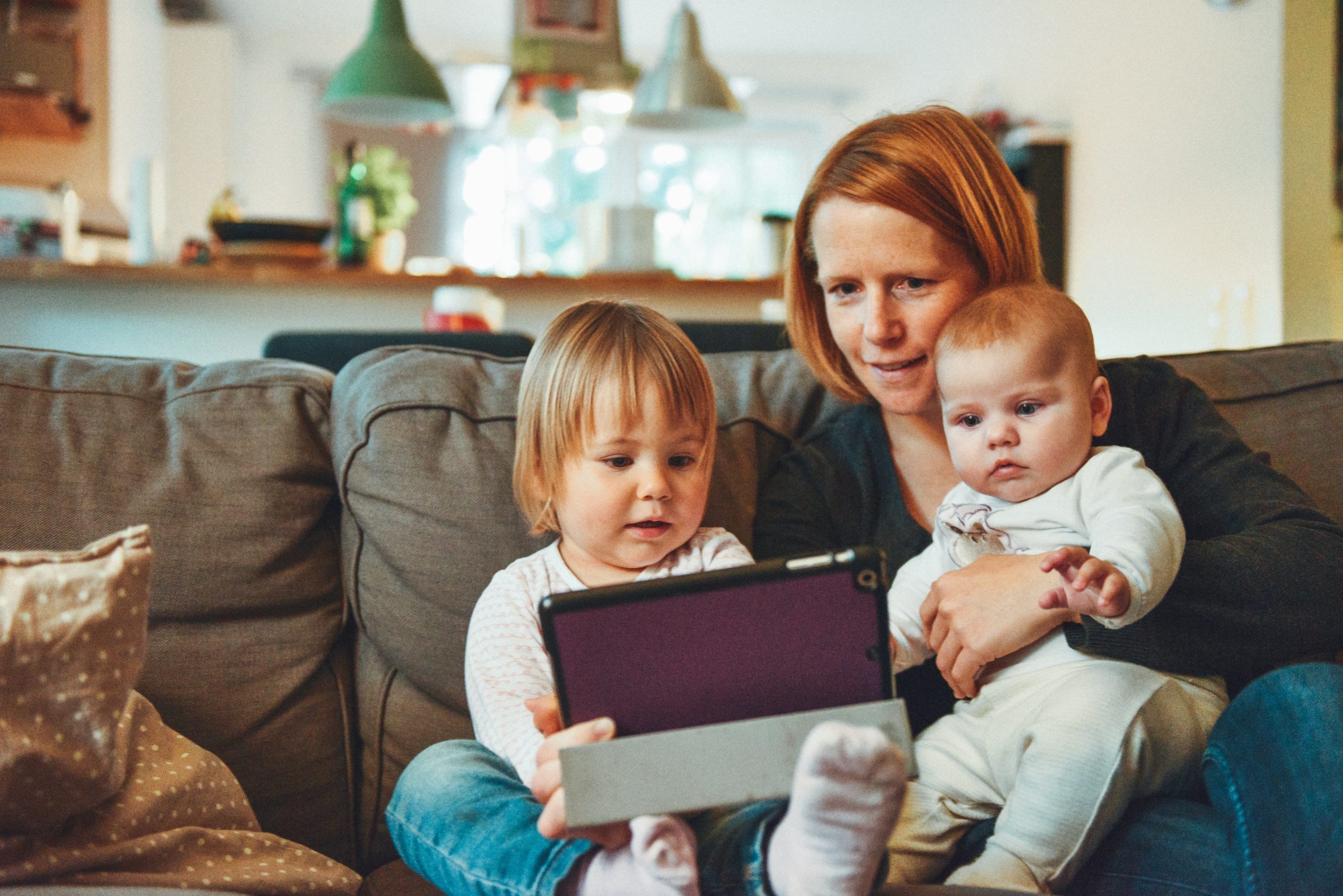 A woman holds a baby on a couch while a toddler sitting next to her holds a tablet. All of them are looking at the tablet, perhaps for a video chat.