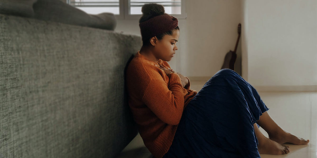 Woman sits on floor at home, leaning on couch, hands on chest, anxious