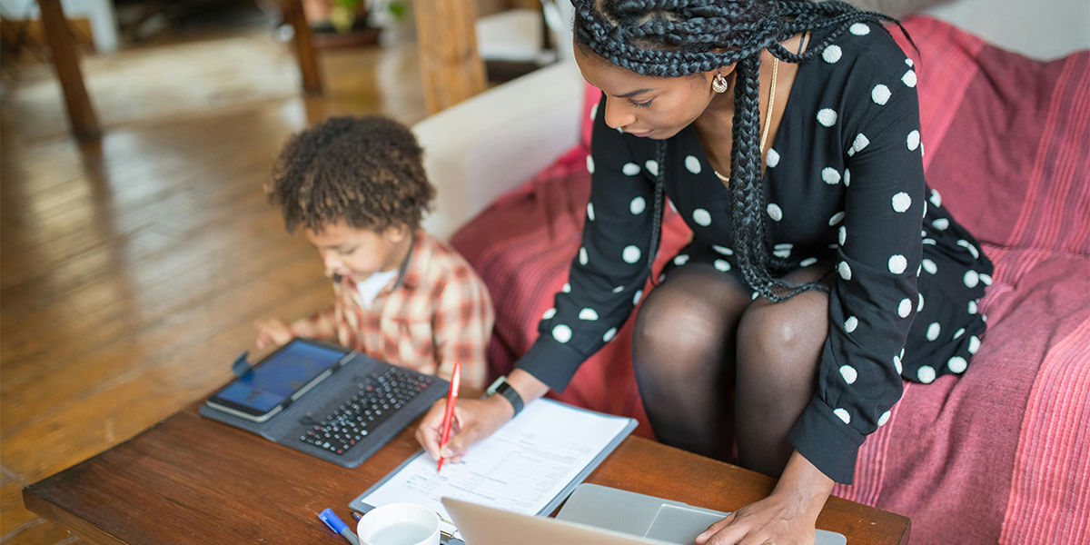 Young American Women Experience Triple Whammy: Financial, Physical and Mental Well-Being Challenges. Mother and child sitting at coffee table, she's working, he's playing on tablet
