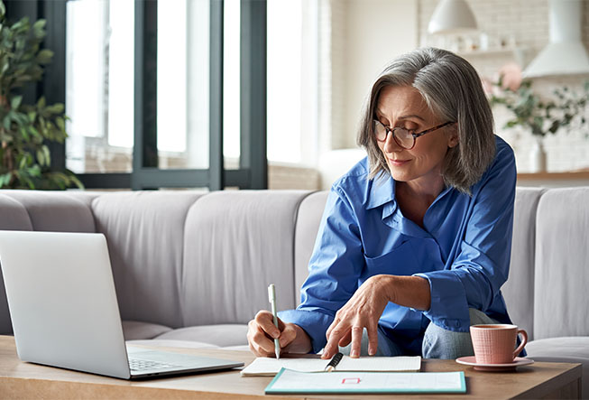 Older woman taking notes while sitting on couch, laptop in front of her