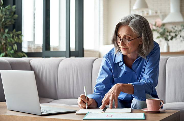 Older woman taking notes while sitting on couch, laptop in front of her