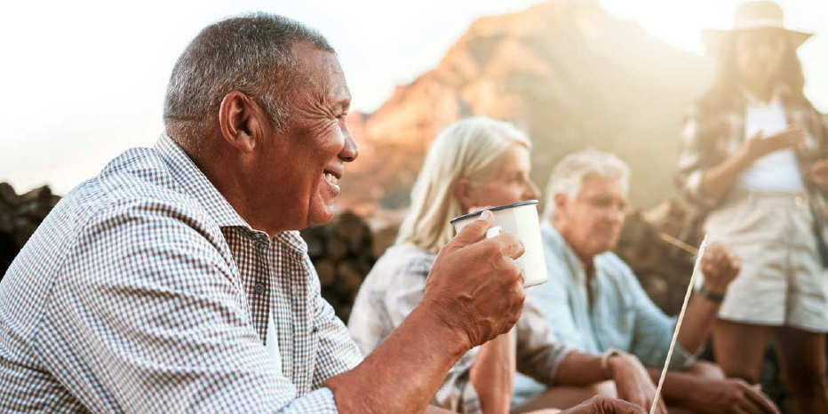 An older man sits outside with a mug in hand, a few other mature people near him